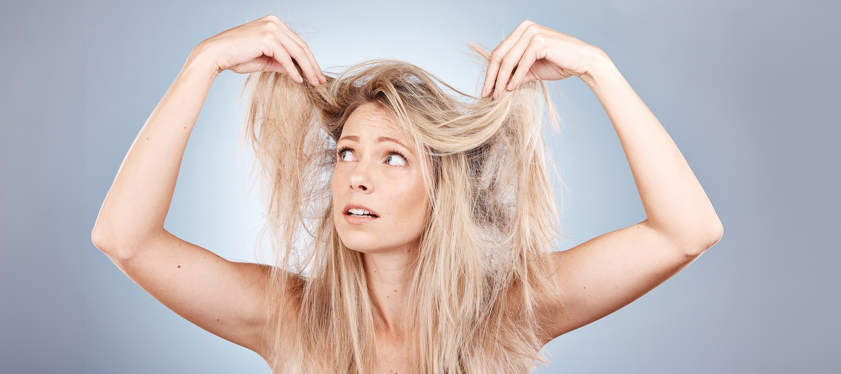 Blonde woman holding up her hair and frustrated with the frizz, against a grey background